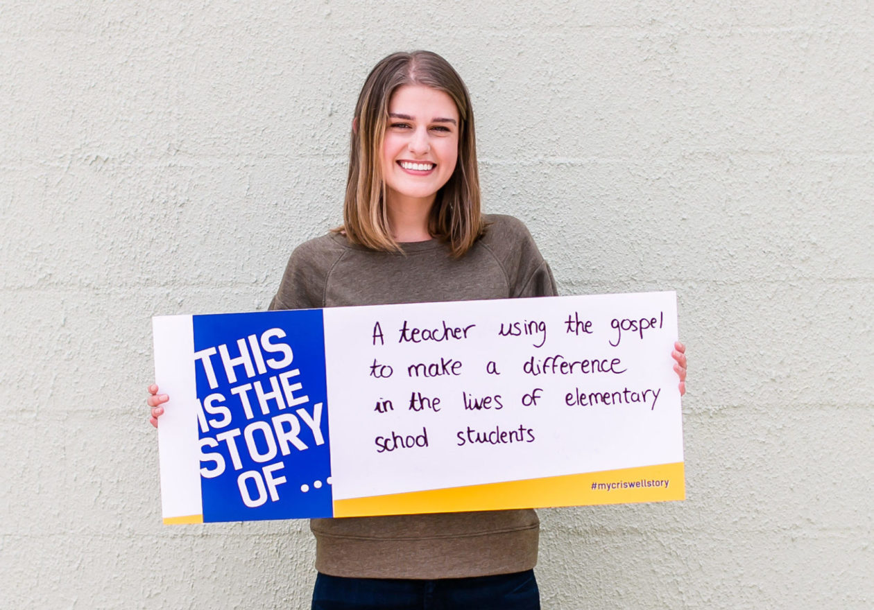 A female undergraduate displaying a sign that states, "This is the story of a teacher using the gospel to make a difference in the lives of elementary school students."