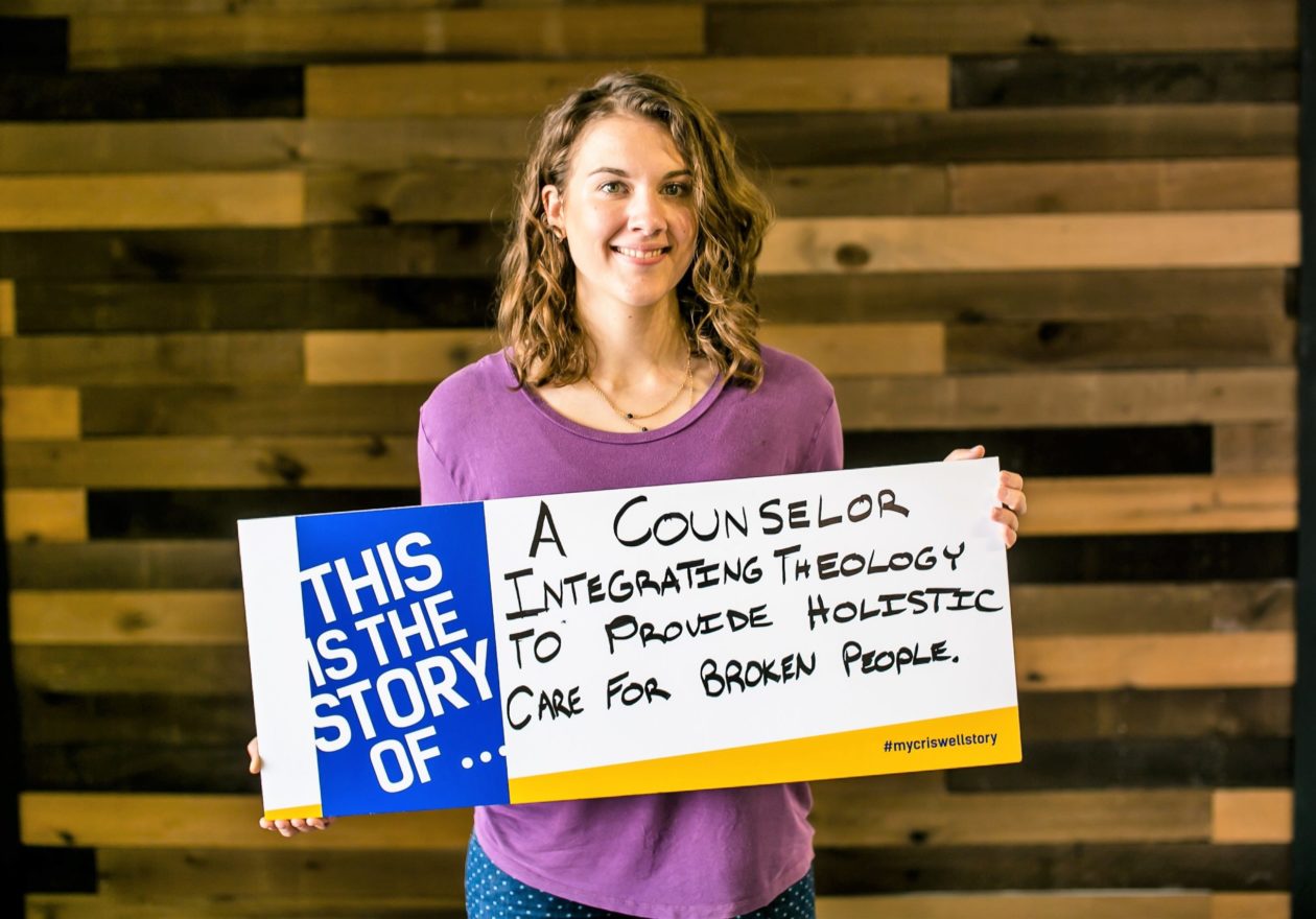 A smiling graduate student displaying a sign that states, "This is the story of a counselor integrating theology to provide holistic care for broken people."