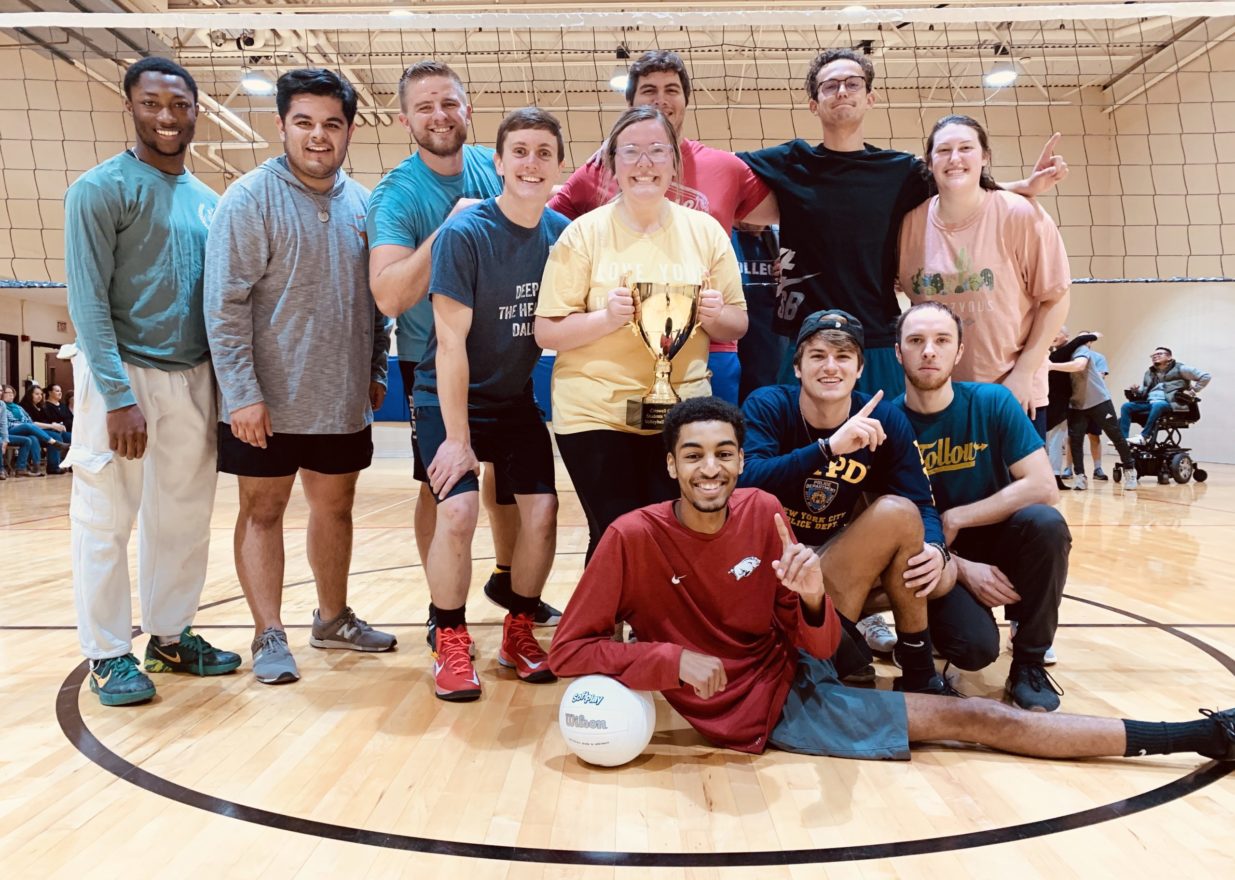 Smiling victorious students holding a volleyball and trophy after beating faculty and staff in a friendly tournament.